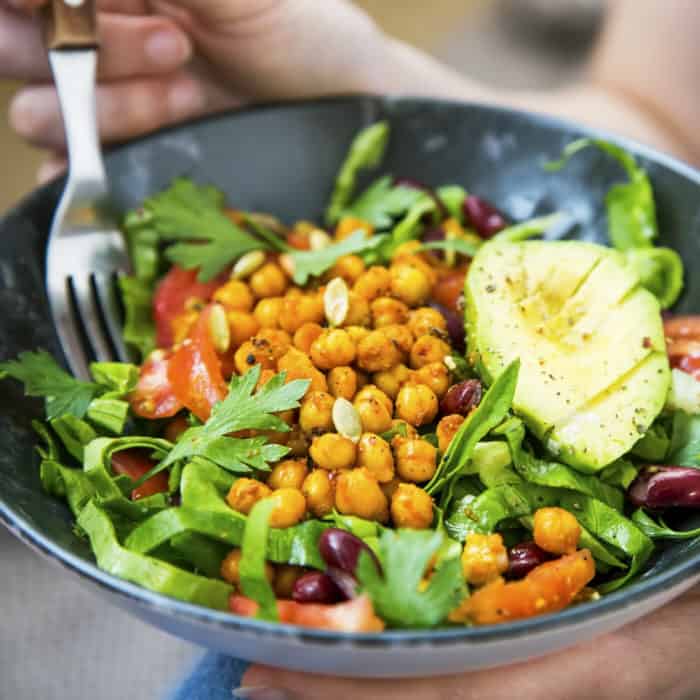 Fork digging into a buddha bowl full of fresh greens, tomatoes, chickpeas, olives and avocado