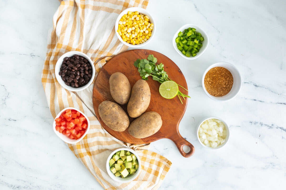 Ingredients for taco baked potatoes including russet potatoes, black beans, corn, diced tomatoes, avocado, onion, cilantro, and lime arranged on a cutting board.