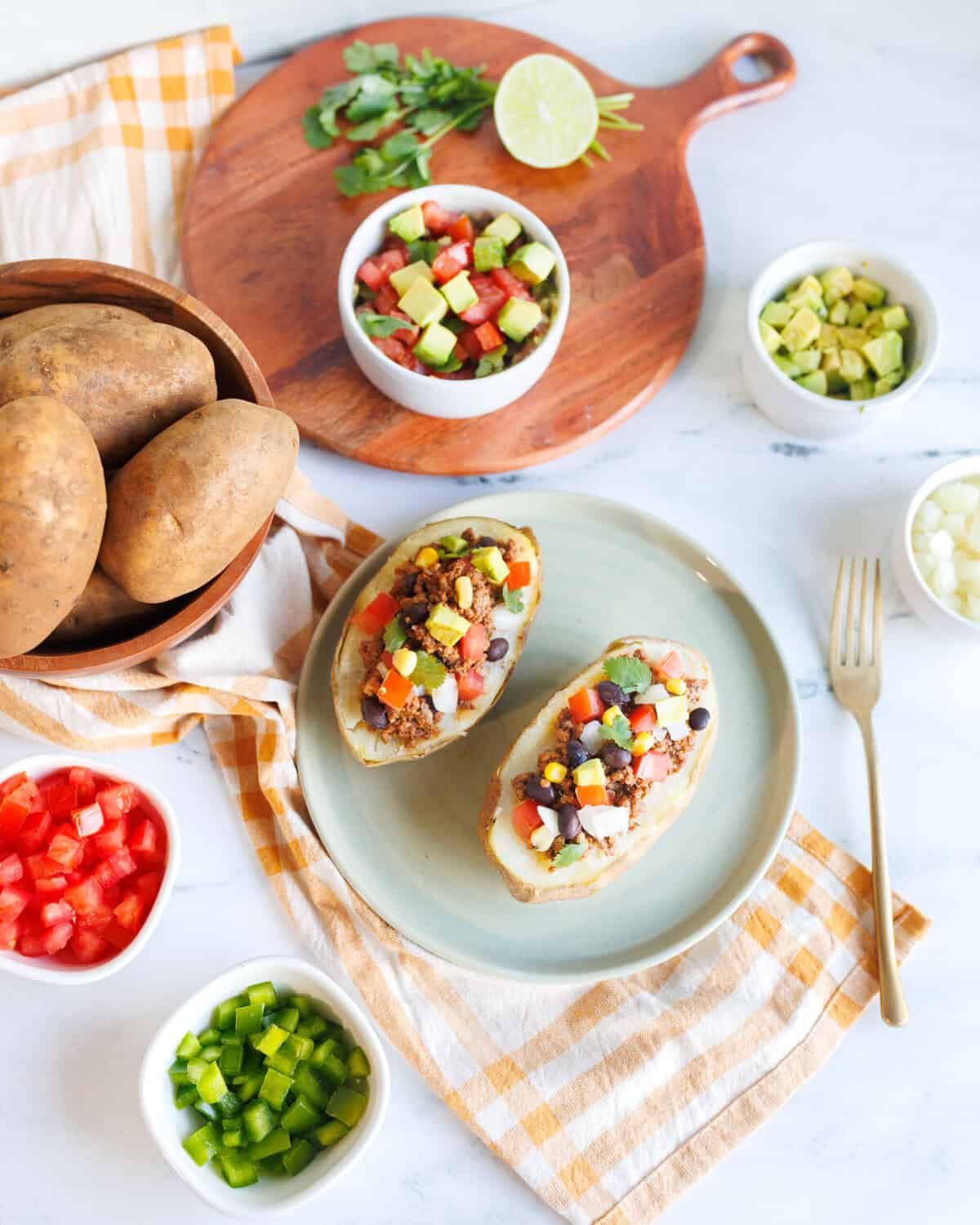 Fiesta Taco Baked Potatoes Loaded taco baked potatoes on a plate surrounded by toppings like diced tomatoes, avocado, black beans, corn, and cilantro for an easy dinner idea.
