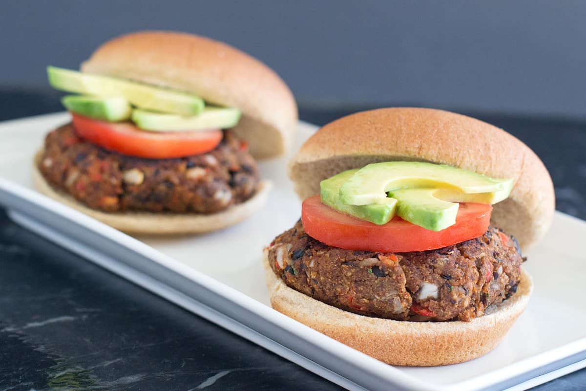 Two black bean burgers topped with tomato and avocado slices and plated on rectangle white plate