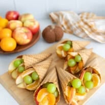 A wooden board topped with waffle cone cornucopias filled with apples, clementine segments, and green grapes, with fresh fruit and a kitchen towel in the background.