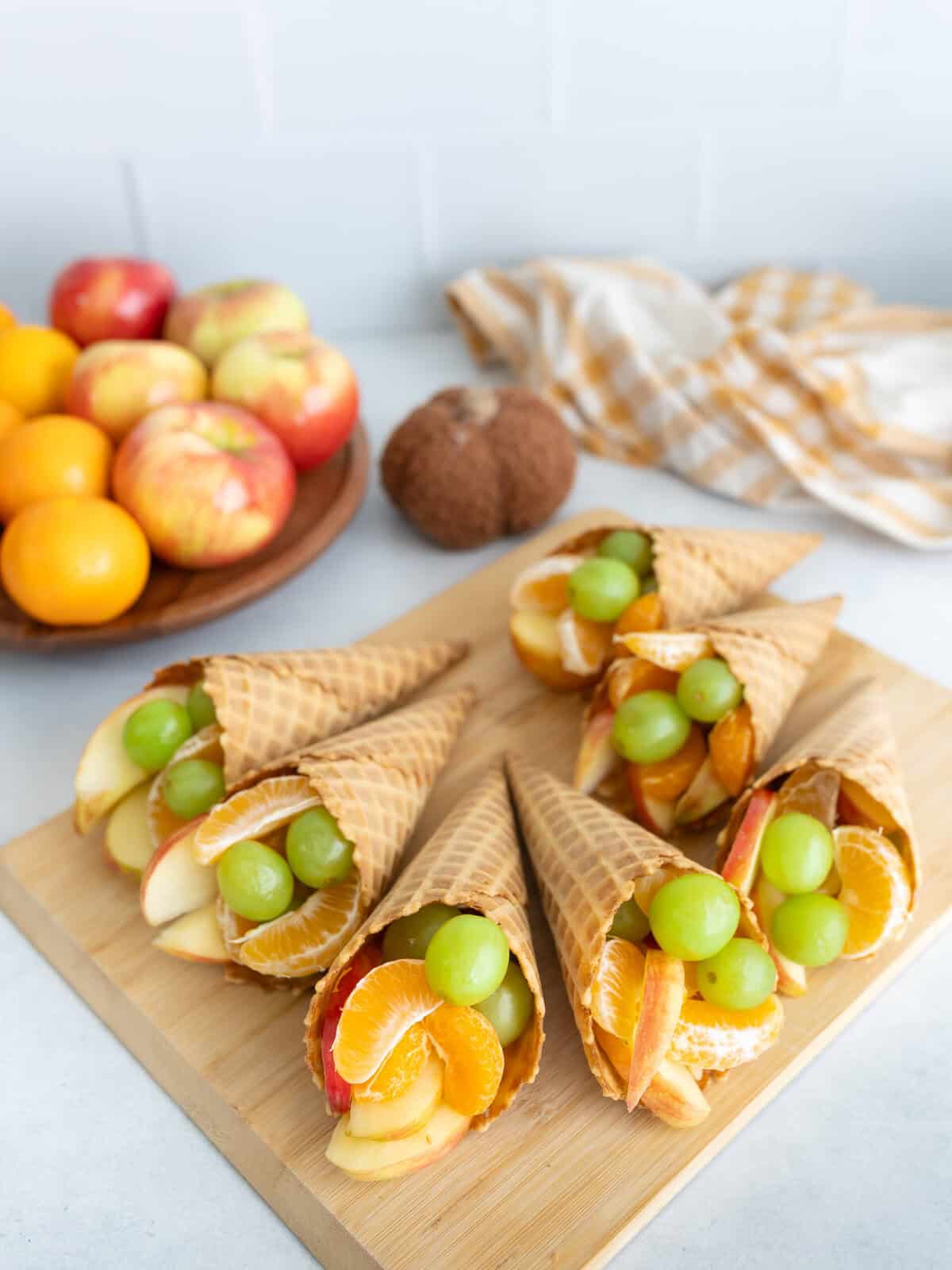Thanksgiving Fruit Cornucopias A wooden board topped with waffle cone cornucopias filled with apples, clementine segments, and green grapes, with fresh fruit and a kitchen towel in the background.