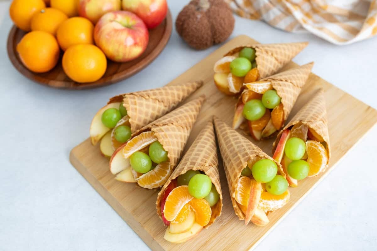 Fruit Cornucopias A wooden board topped with waffle cone cornucopias filled with apples, clementine segments, and green grapes, with fresh fruit and a kitchen towel in the background.