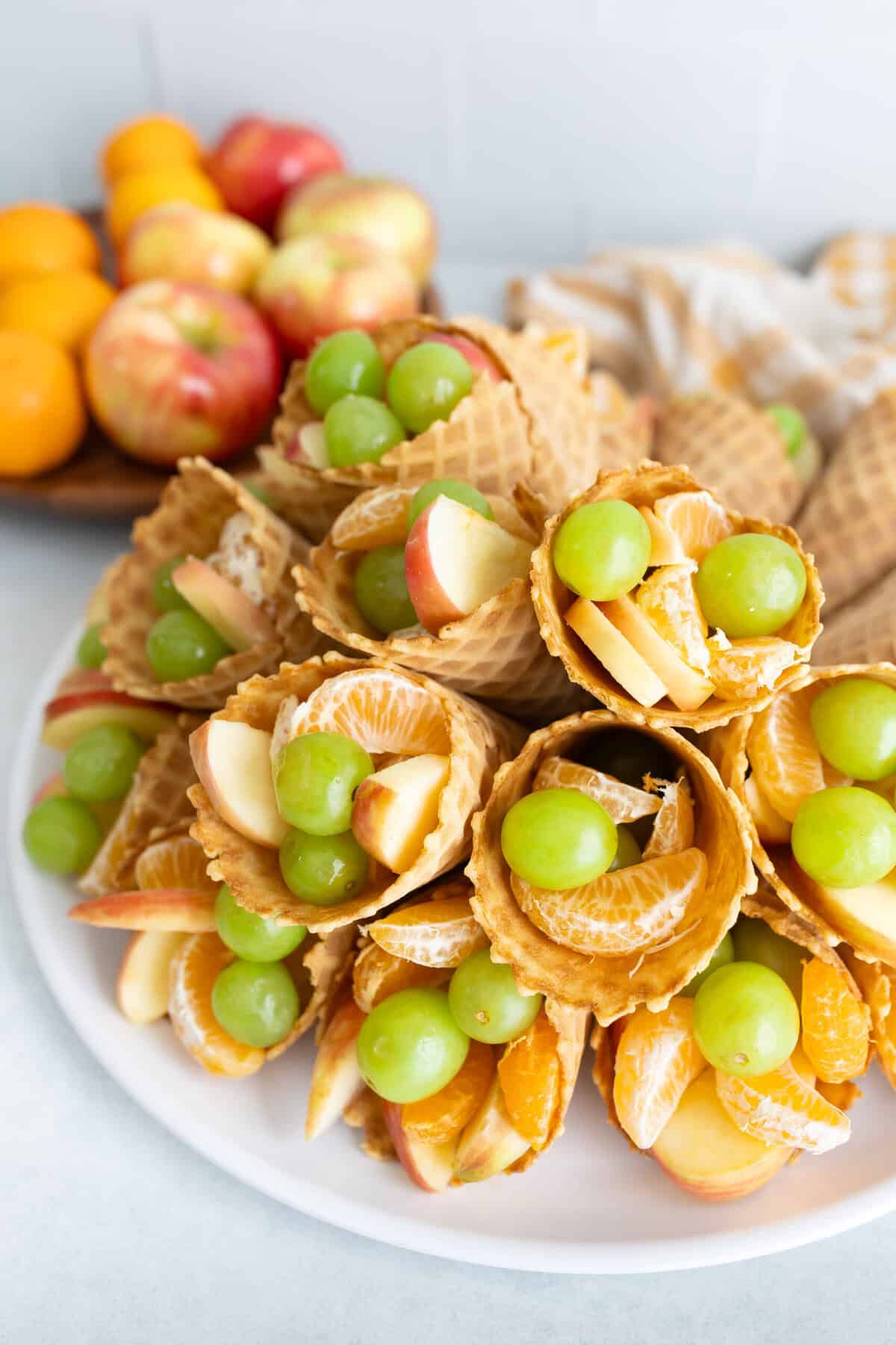 how to make thanksgiving fruit cornucopias A wooden board topped with waffle cone cornucopias filled with apples, clementine segments, and green grapes, with fresh fruit and a kitchen towel in the background.