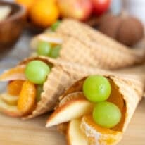 A wooden board topped with waffle cone cornucopias filled with apples, clementine segments, and green grapes, with fresh fruit and a kitchen towel in the background.