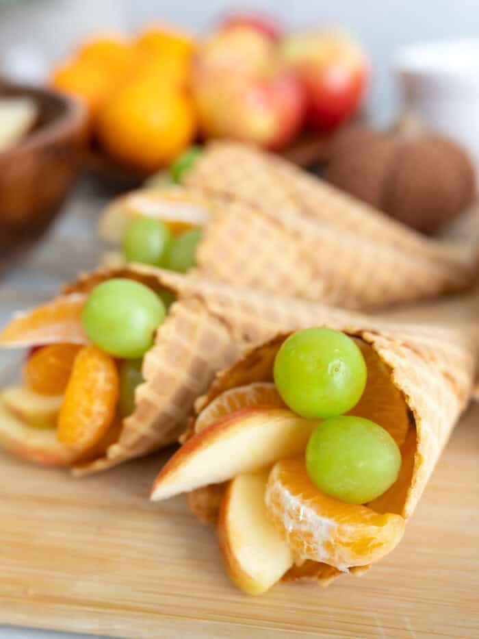 A wooden board topped with waffle cone cornucopias filled with apples, clementine segments, and green grapes, with fresh fruit and a kitchen towel in the background.