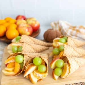 A wooden board topped with waffle cone cornucopias filled with apples, clementine segments, and green grapes, with fresh fruit and a kitchen towel in the background.