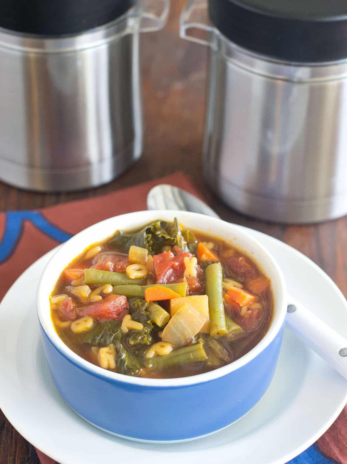 Vegetable soup in blue bowl on white plate with spoon. Two thermos containers shown in background