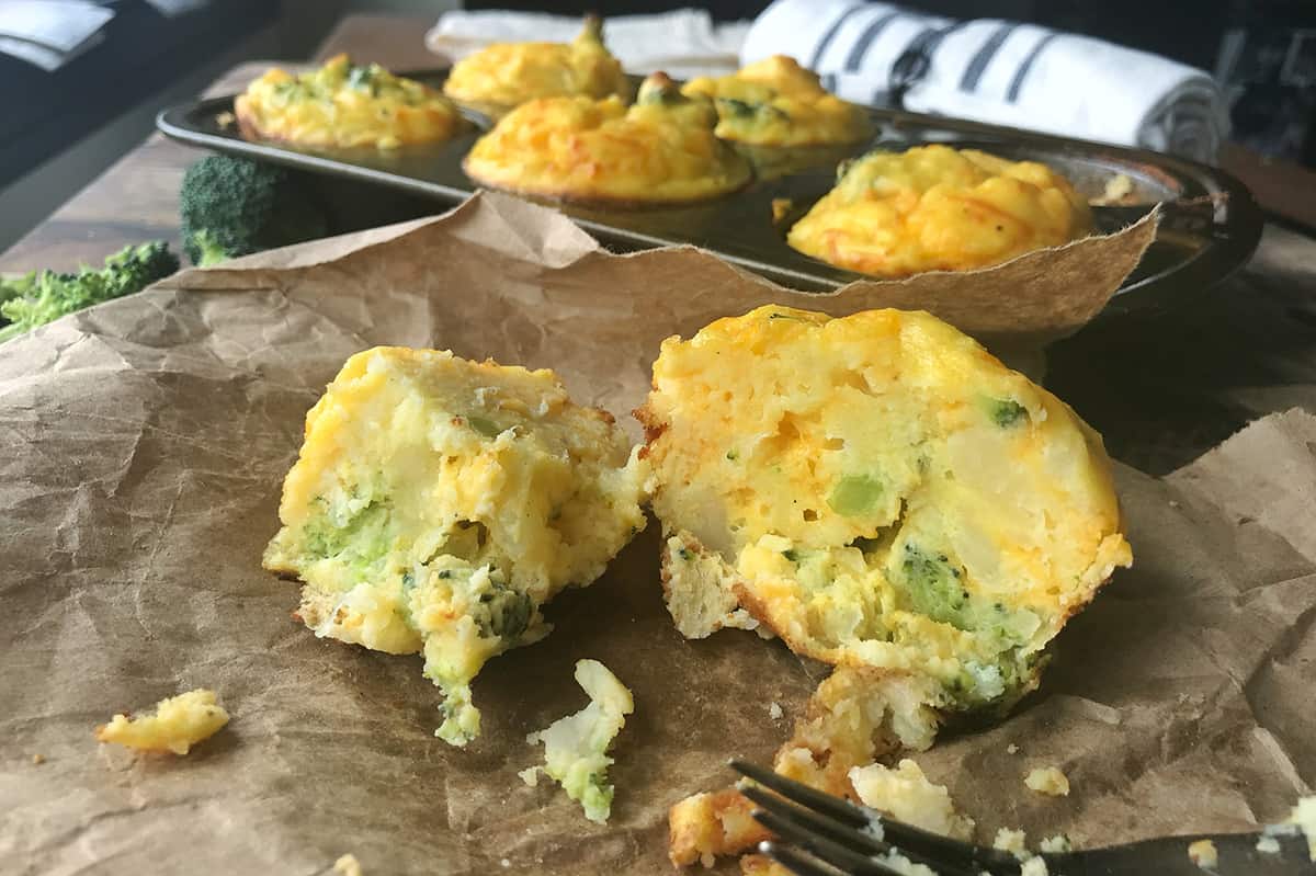 Mashed potato bite broken open on parchment paper. Muffin tray of additional mashed potato bites in the background