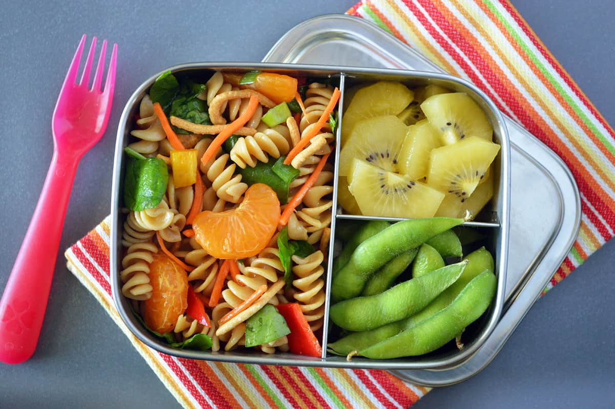Metal bento box filled with pasta salad, kiwi and edamame on striped napkin and a pink plastic fork
