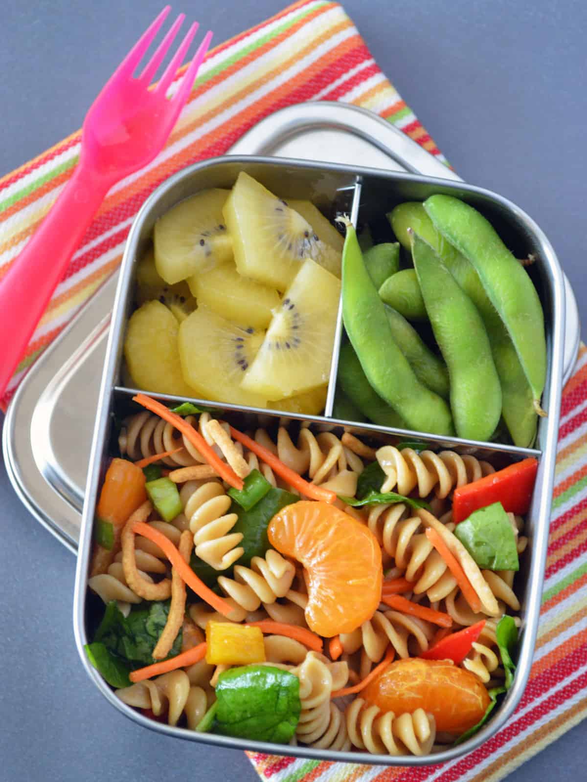 Metal bento box filled with pasta salad, kiwi and edamame on striped napkin and a pink plastic fork