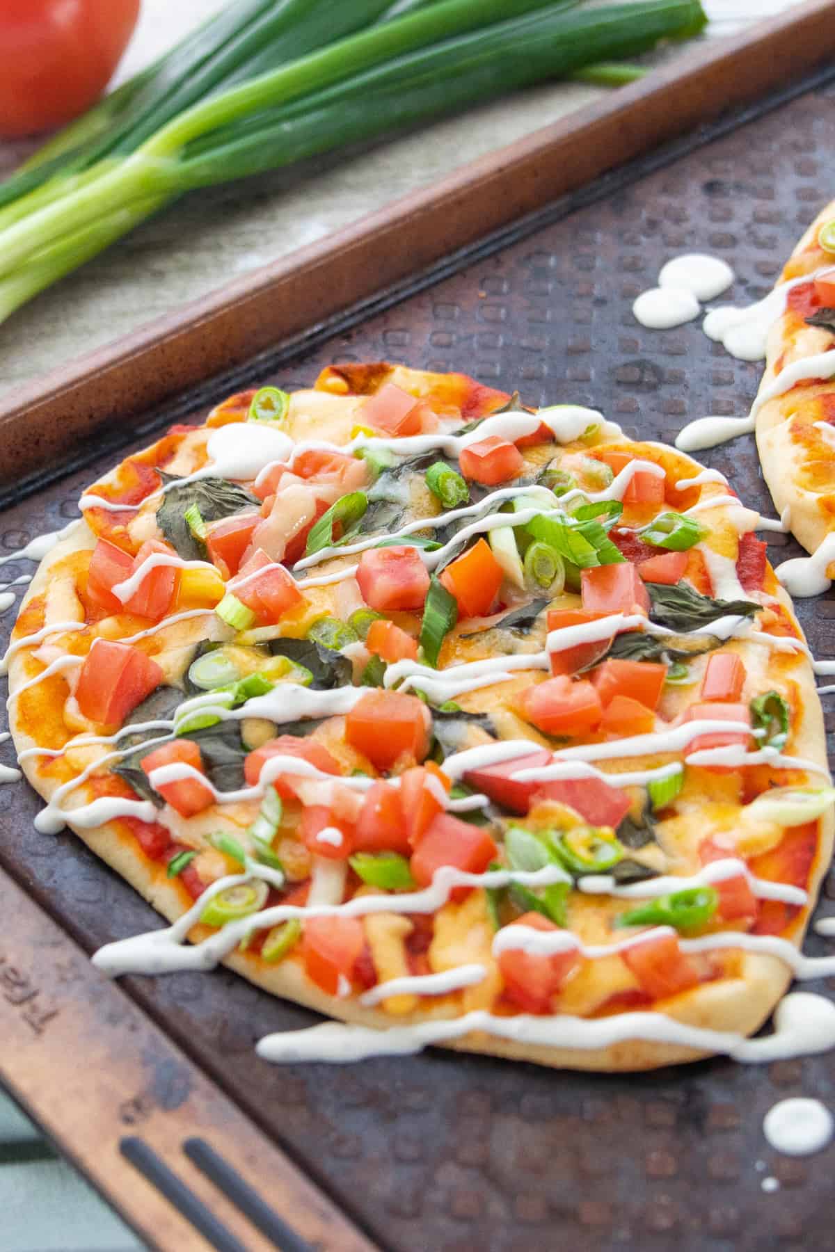 Naan flatbread pizzas topped with diced tomatoes, sliced green onions, leafy greens, melted cheese, and a creamy drizzle, served on a baking sheet with fresh green onions and a whole tomato nearby.