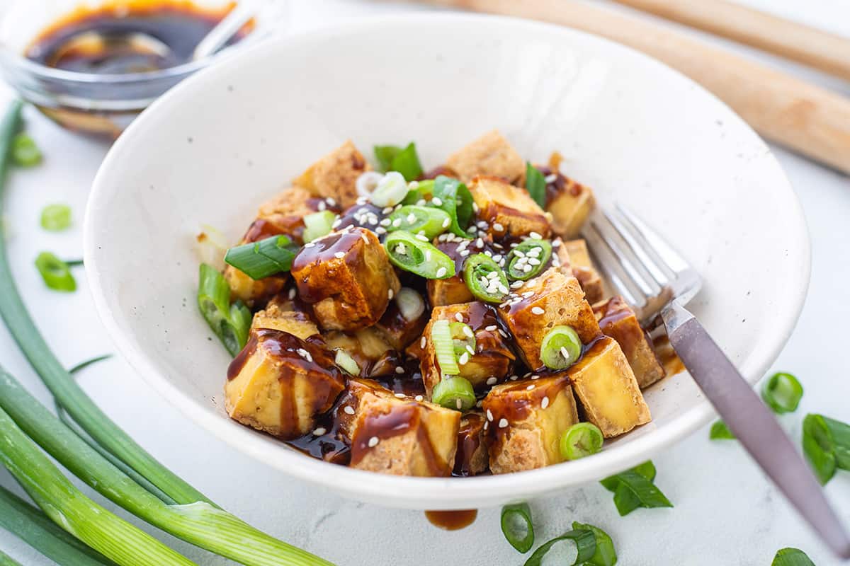 White bowl with crispy tofu drizzled with sauce and sprinkled with green onions and sesame seeds, plated with a fork.