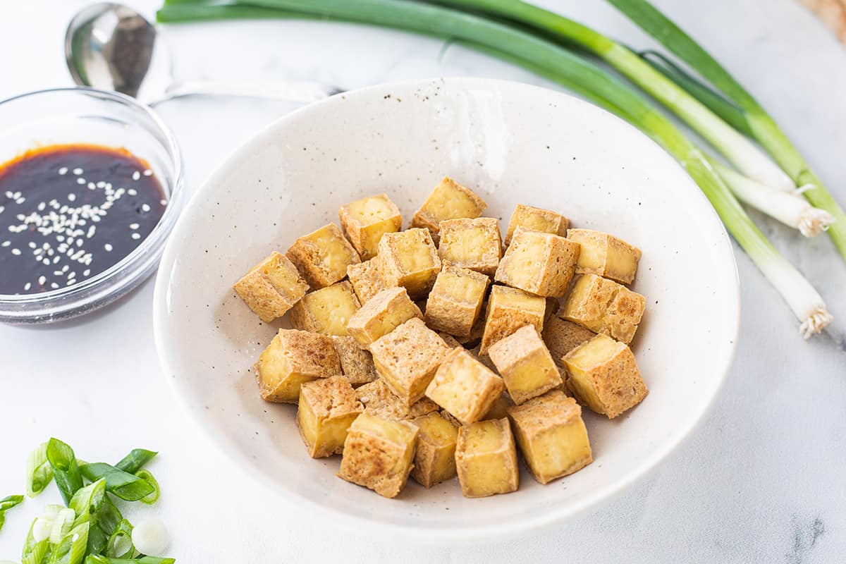 Crispy baked tofu in white bowl with whole green onions and a small bowl of sauce in background