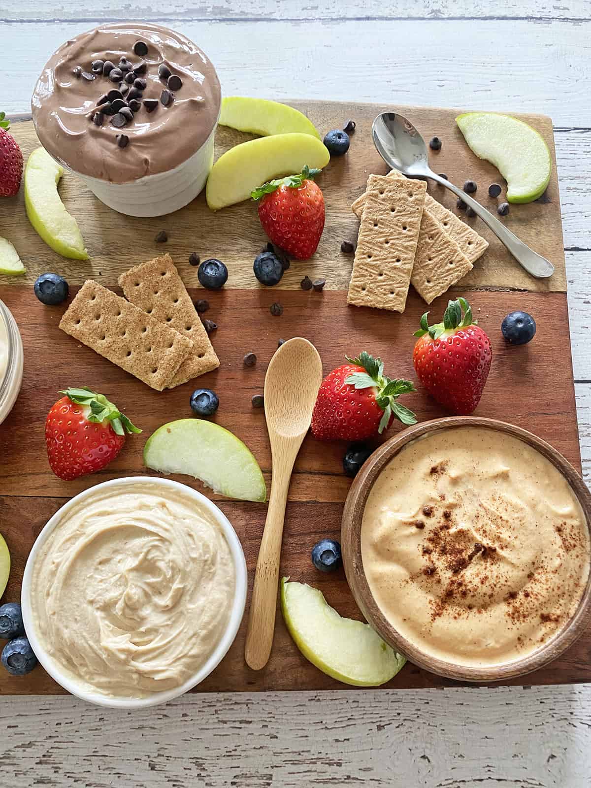 Close up of snack board with fruit dips in bowls, sliced apples, strawberries, blueberries and graham crackers.