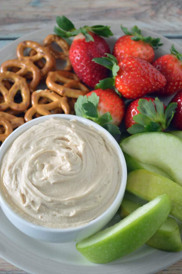 Bowl of peanut butter yogurt dip plated with sliced apples, strawberries and pretzels.