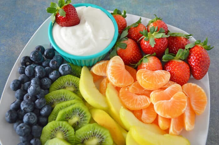 Cream cheese fruit dip in bowl plated with blueberries, kiwi, yellow apples, mandarins and strawberries.