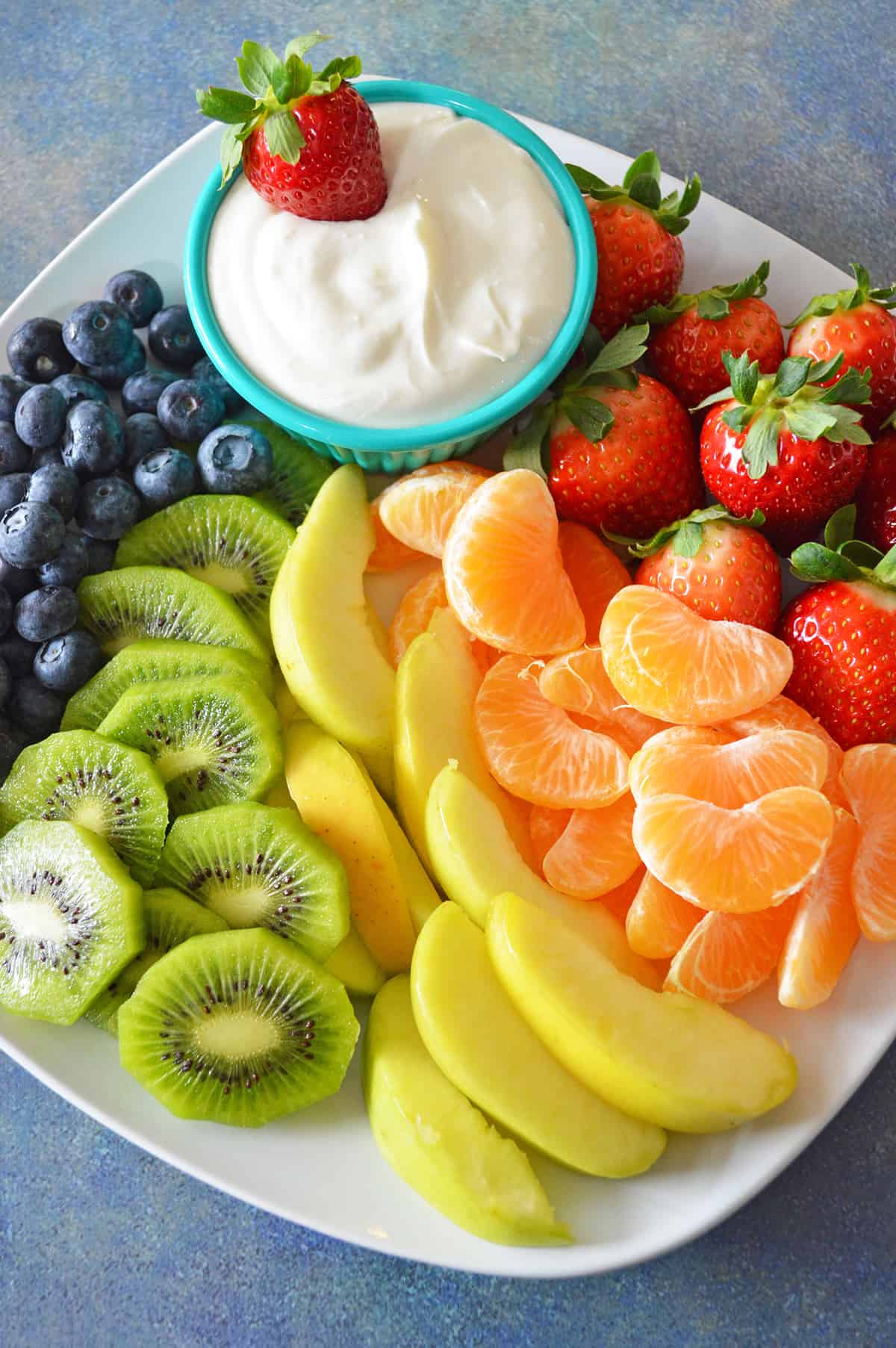 Cream cheese fruit dip in bowl plated with blueberries, kiwi, yellow apples, mandarins and strawberries.