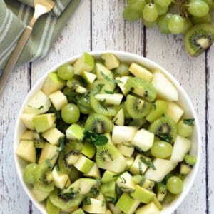 Green fruit salad in white back on whitewashed wood table. Napkin, fork and green grapes in background.