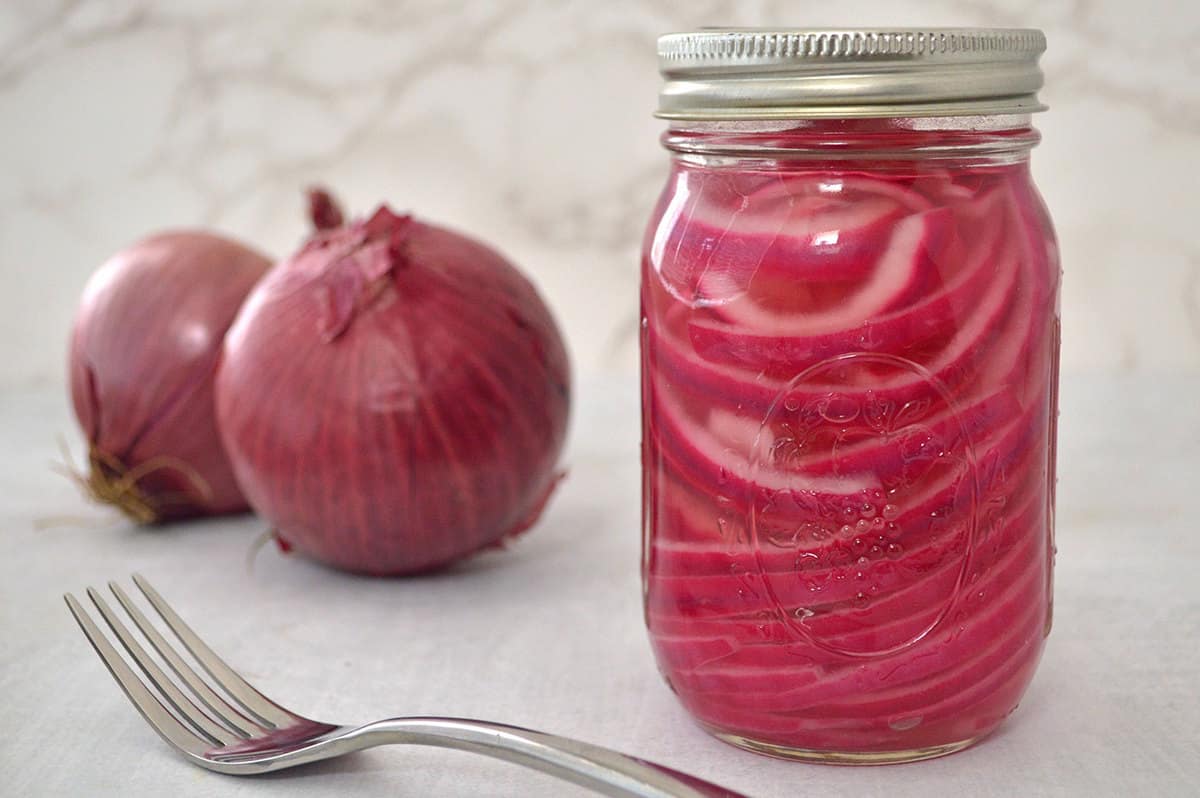 Mason jar filled with pickled red onions with a fork in front. Two large whole red onions in the background.