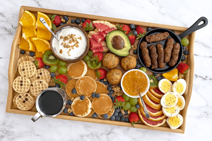 Overhead of breakfast grazing board filled with mini waffles, pancakes, fruit, hard-boiled eggs, sausage links, yogurt and more.