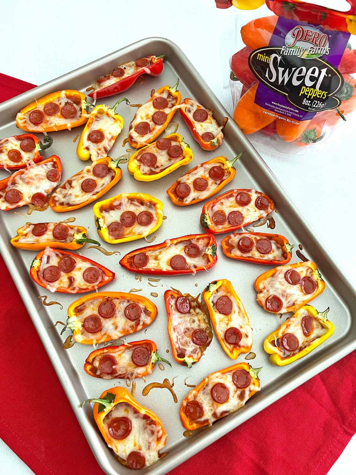 Cooked mini pepper pizzas on baking sheet on top of red towel with package of mini sweet peppers in background.