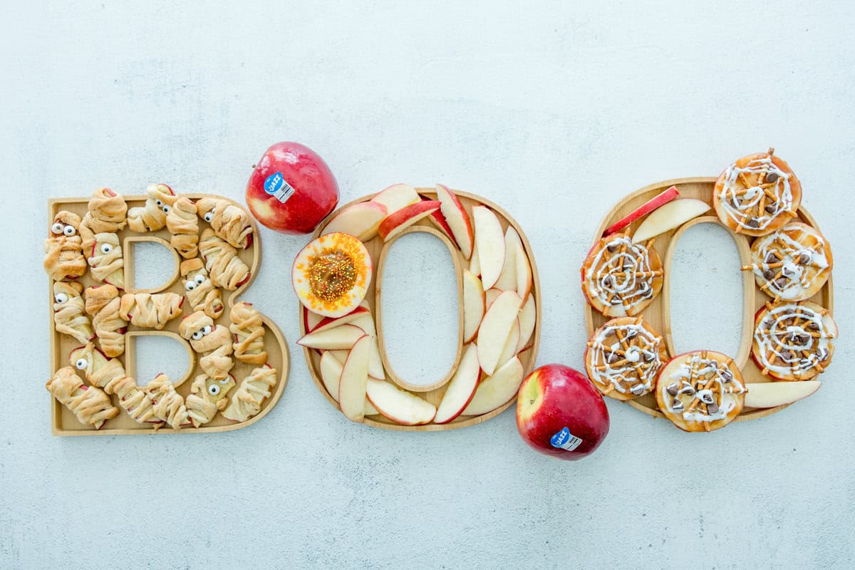 Halloween treats board spelling BOO with apple slices, cinnamon rolls, and mummies.