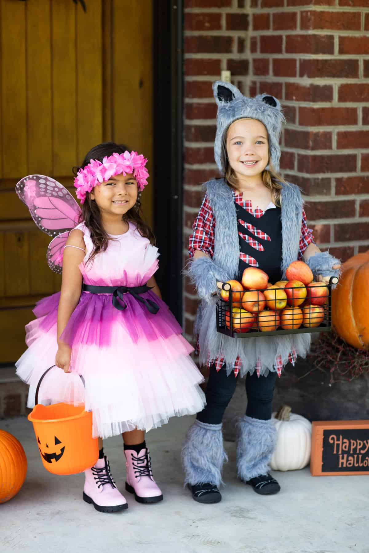 Two young girls dressed in costumes. One is a butterfly fairy, and the other is a wolf carrying a basket of apples.