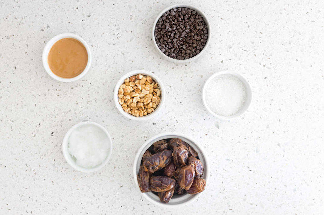 Bowls with ingredients needed for preparing Date Bark: chocolate chips, peanuts, sea salt, coconut oil, peanut butter, and dates.