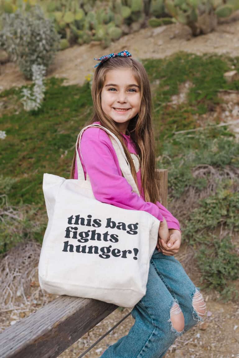 A little girl sitting over a fence holding a tote bag