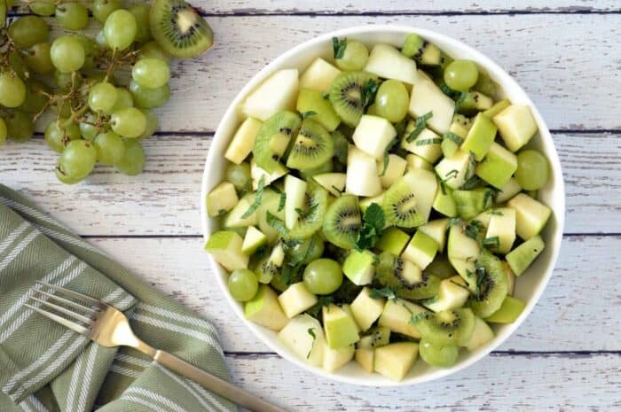 Green fruit salad in white back on whitewashed wood table. Napkin, fork and green grapes in background.