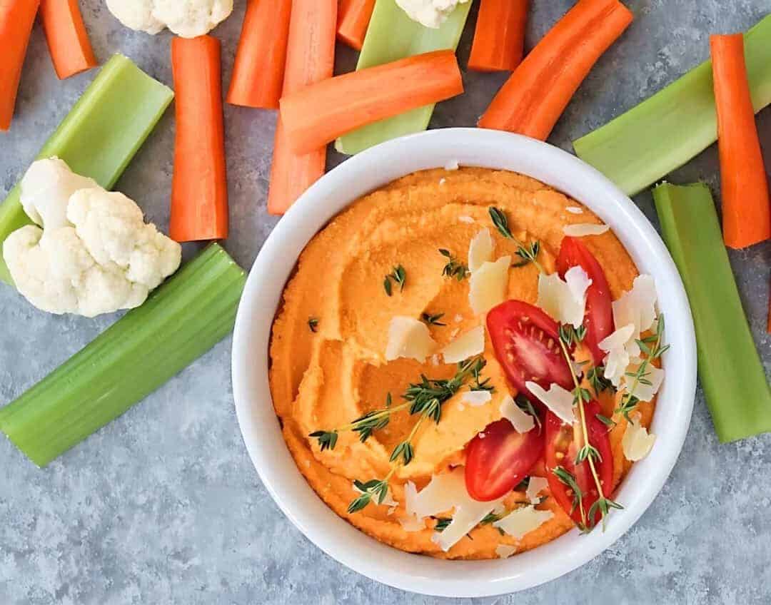 Roasted Tomato Hummus recipe in a white bowl, garnished with thyme and paired with celery and carrot sticks.