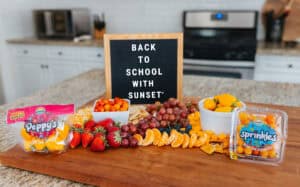 Back-to-school snack board on a kitchen island featuring Sunset&reg; Sprinkles tomatoes, Peppy&rsquo;s teeny tiny peppers, fruit, cheese cubes, and a letterboard sign reading &ldquo;Back to School with SUNSET&reg;.&rdquo;