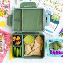 Open green lunchbox on a school-themed background with mini cucumbers, tortilla wraps, guacamole, mandarin slices, blueberries, and Horizon Organic chocolate milk. Nature Fresh Farms Lil Chills™ mini cucumbers are featured alongside school supplies and a back-to-school book.