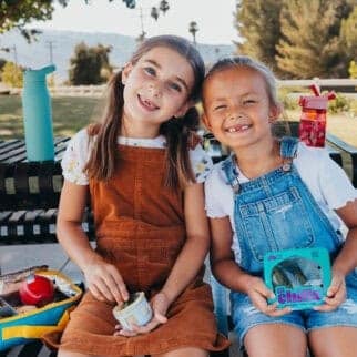 Two smiling young girls sitting at a park bench enjoying lunch, one holding a cup of dip and the other holding a package of Nature Fresh Farms Lil Chills™ mini cucumbers — a fun and healthy back-to-school snack option.