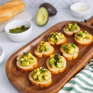 Avocado bruschetta appetizer on a wooden platter with fresh avocado, baguette slices, and herb oil in the background.