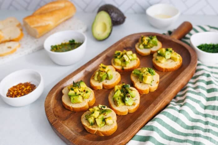 Avocado bruschetta appetizer on a wooden platter with fresh avocado, baguette slices, and herb oil in the background.