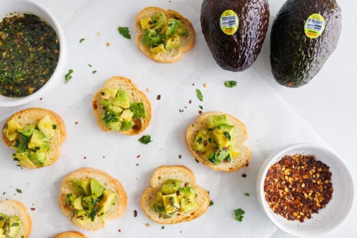 Overhead view of avocado bruschetta bites with whole avocados, herb oil, and red pepper flakes on a white surface.
