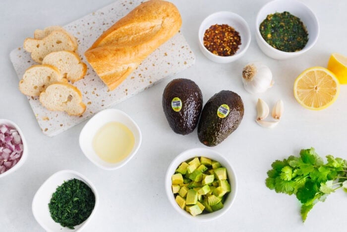 Ingredients for avocado bruschetta including fresh avocados, baguette, lemon, garlic, herbs, and olive oil.
