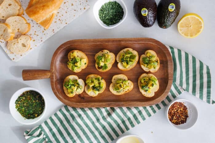 Avocado bruschetta arranged on a wooden serving board with sliced baguette, herbs, and lemon.