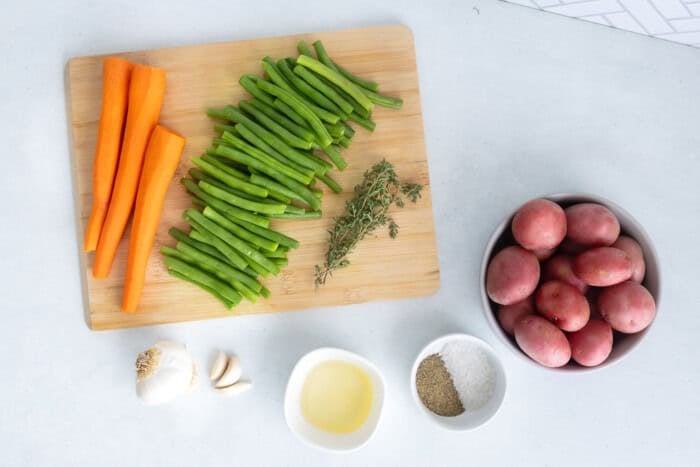 Ingredients for roasted baby potatoes with green beans and carrots including garlic, olive oil, and fresh herbs